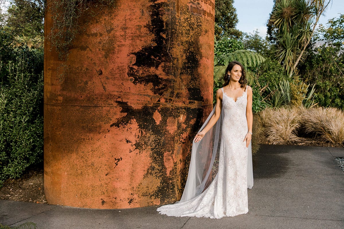 Model wearing Vinka Design Arabella Wedding Dress, a Beaded Lace Wedding Gown with Cape in Hamilton Gardens landscape