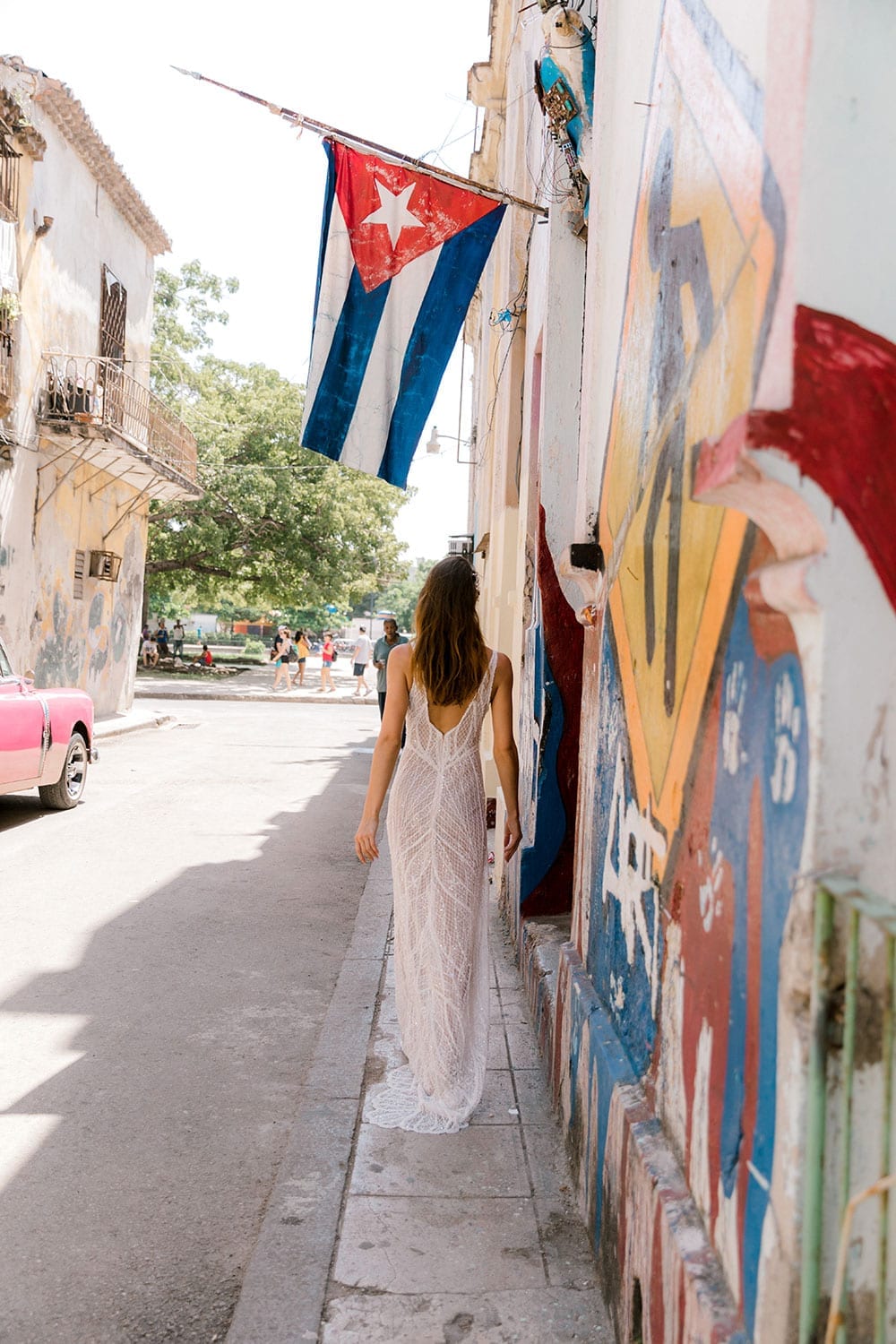 Model wearing Vinka Design Alexia Wedding Dress, a Semi Sheer Beaded Lace Wedding Gown walking away on Havana street with flag and roadsign