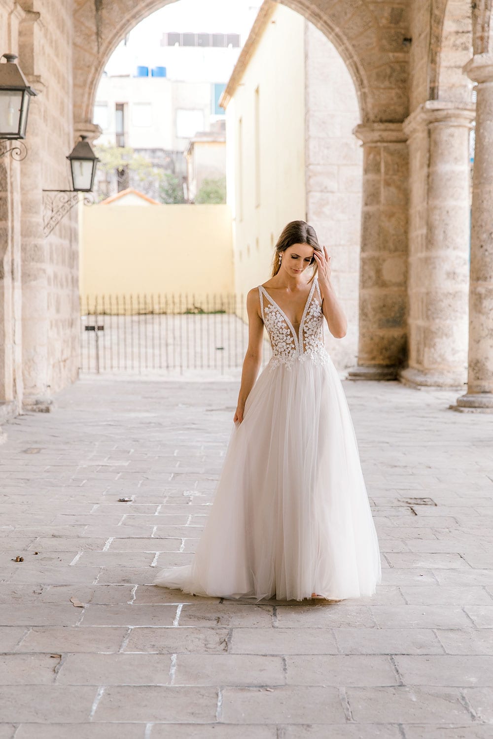 Model wearing Vinka Design Isabel Wedding Dress, a Champagne coloured Tulle Gown with Sheer V-Neck Bodice and Beaded Lace in the arch of an old building in Havana