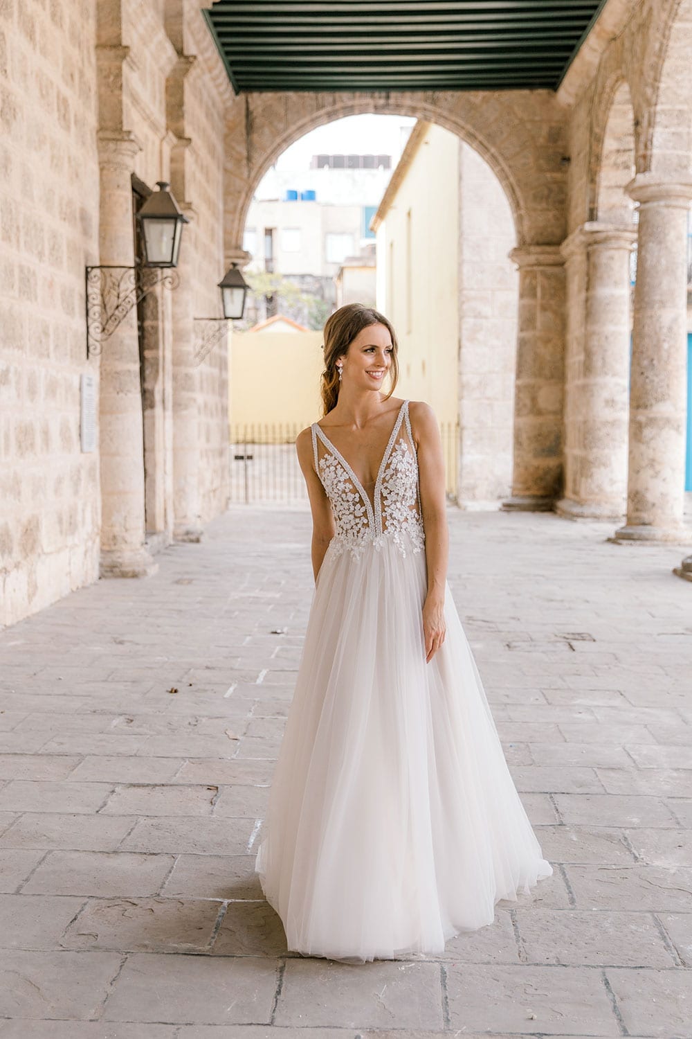 Model wearing Vinka Design Isabel Wedding Dress, a Champagne coloured Tulle Gown with Sheer V-Neck Bodice and Beaded Lace in the arches of an old building in Havana portrait