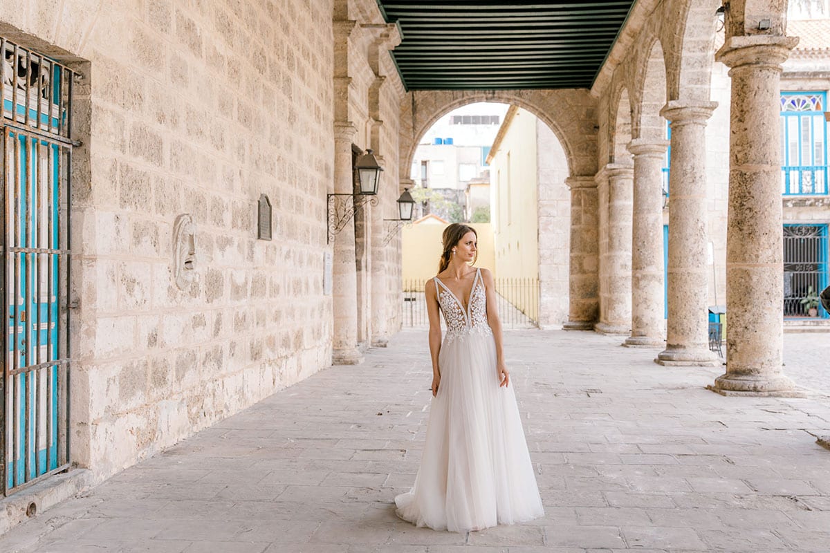 Model wearing Vinka Design Isabel Wedding Dress, a Champagne coloured Tulle Gown with Sheer V-Neck Bodice and Beaded Lace in the arches of an old building in Havana