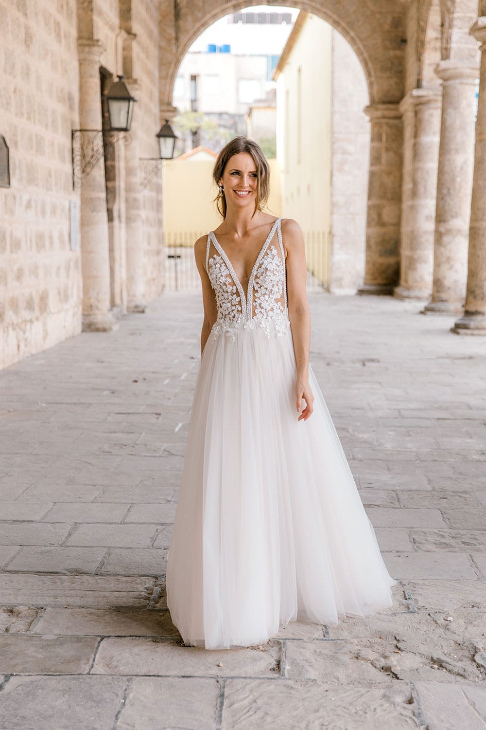 Model wearing Vinka Design Isabel Wedding Dress, a Champagne coloured Tulle Gown with Sheer V-Neck Bodice and Beaded Lace in the archway of an old building in Havana