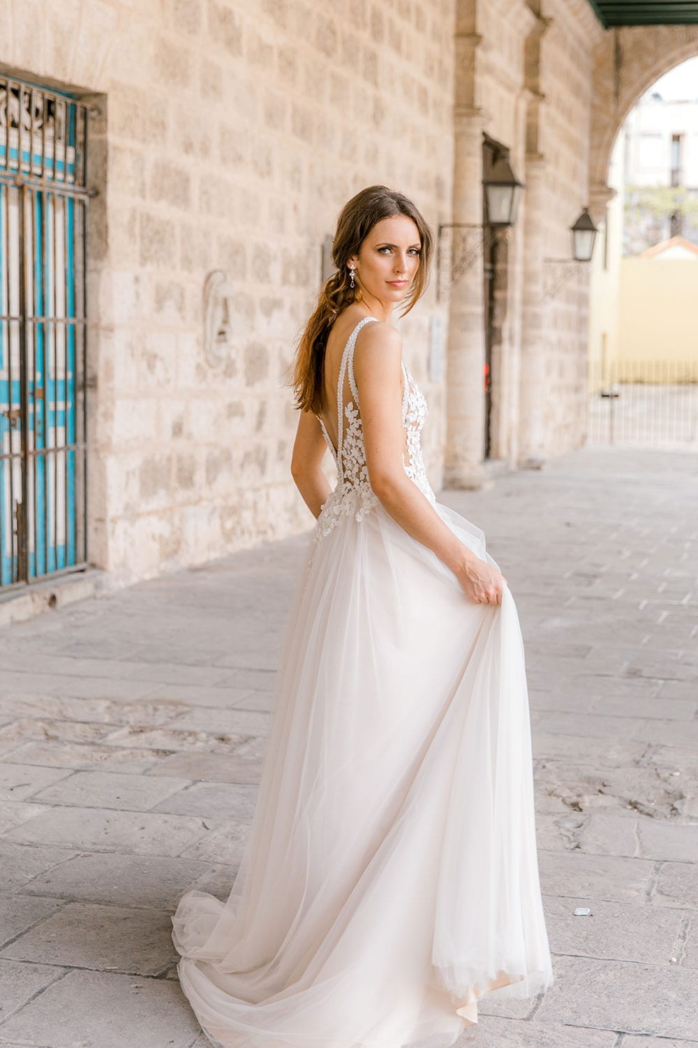 Model wearing Vinka Design Isabel Wedding Dress, a Champagne coloured Tulle Gown with Sheer V-Neck Bodice and Beaded Lace in the archway of an old building in Havana facing away