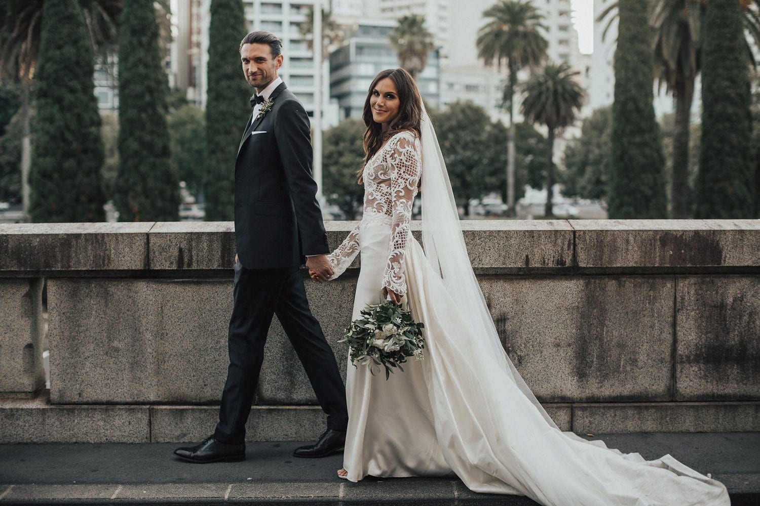 Real Weddings | Vinka Design | Real Brides Wearing Vinka Gowns | Hannah and Campbell - walking along bridge dress in light showing lace detail, train and veil