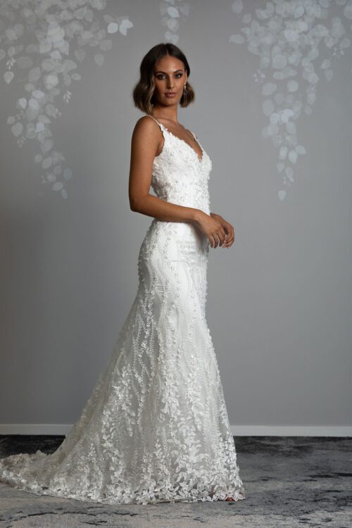 Camille Wedding Dress by Vinka Design. Timeless fitted gown that is sculpted to the body, featuring a beautiful and delicate three dimensional flower lace. Sheer lace over the shoulder draws the eye to the elegant low back. Profile view of model wearing 3D flower lace bodice with shoestring straps and fitted beaded lace skirt