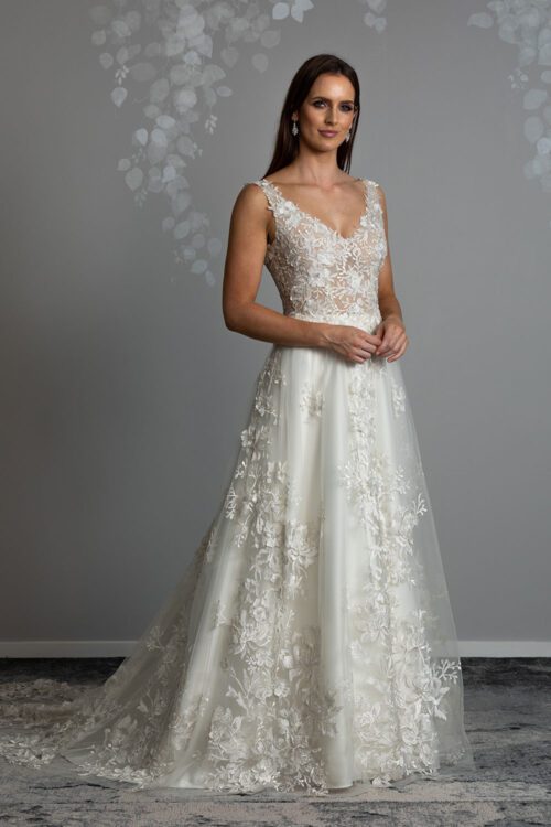 Delta Wedding Dress from Vinka Design. Romantic v-neck gown with a soft silhouette of layers of tulle and delicate 3D lace. The gown's bodice is slightly sheer, creating a nude effect on the skin. Model with hands clasped in front wearing romantic V neck gown with sheer embroidered 3D lace
