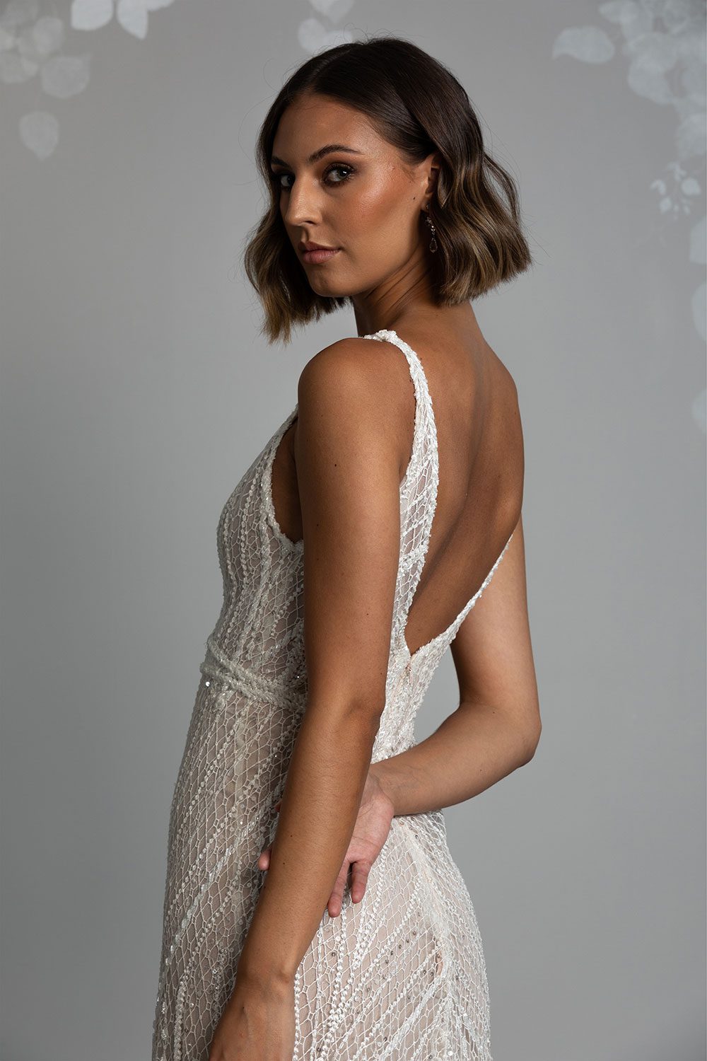 Alexia Wedding Dress by Vinka Design. Featuring a plunging V-neckline and low V-back, this bridal look balances femininity with daring design. Crafted on a nude base, the ivory and silver beads glimmer with movement as they catch light while the lined lace design elongates the female form to perfection. The effortlessly chic sheath skirt falls gently from the waist into a delicate and discreet train. Close up of profile view of model wearing low V back semi sheer beaded lace gown