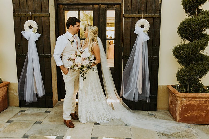 Testimonials | Vinka Design | Real Bridal Reviews from Our Clients | Camilla and Aaron face to face in doorway with bespoke dress placed beautifully along grounds with veil