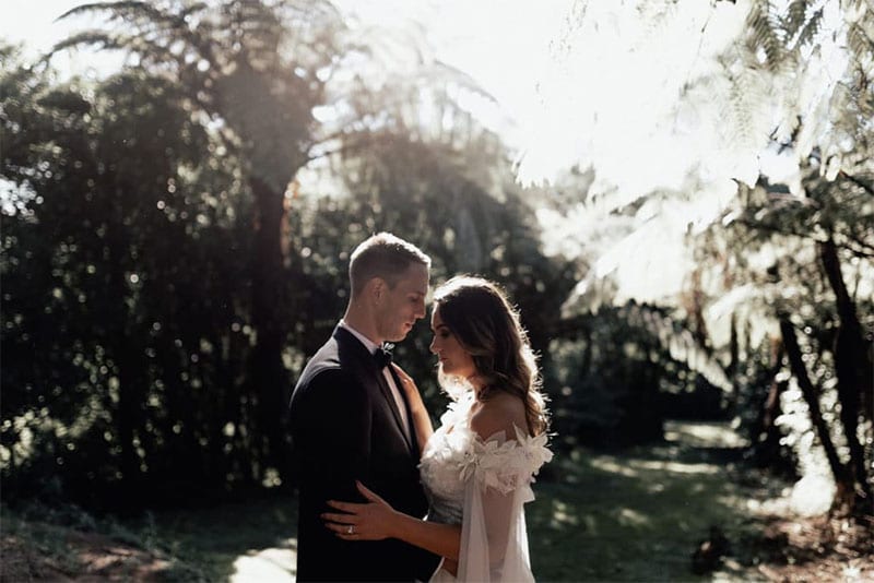 Testimonials | Vinka Design | Real Bridal Reviews from Our Clients | Candice and Michael face to face in dramatic outdoors forest area, the bride's bespoke dress with drop shoulder featuring in the light