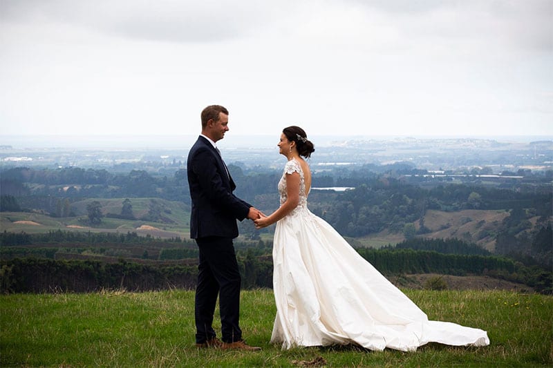 Testimonials | Vinka Design | Real Bridal Reviews from Our Clients | Emmalee and husband atop a hill face to face with stunning bespoke gown train layers showing