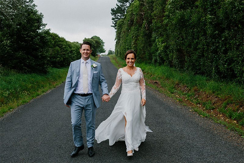 Testimonials | Vinka Design | Real Bridal Reviews from Our Clients | Helen and Mark walking along road holding hands bespoke dress with leg split showing