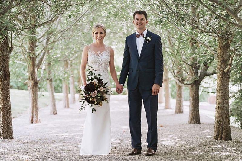 Testimonials | Vinka Design | Real Bridal Reviews from Our Clients | Melissa and husband in tree lined path holding hands, lace detail of bespoke wedding dress highlighted