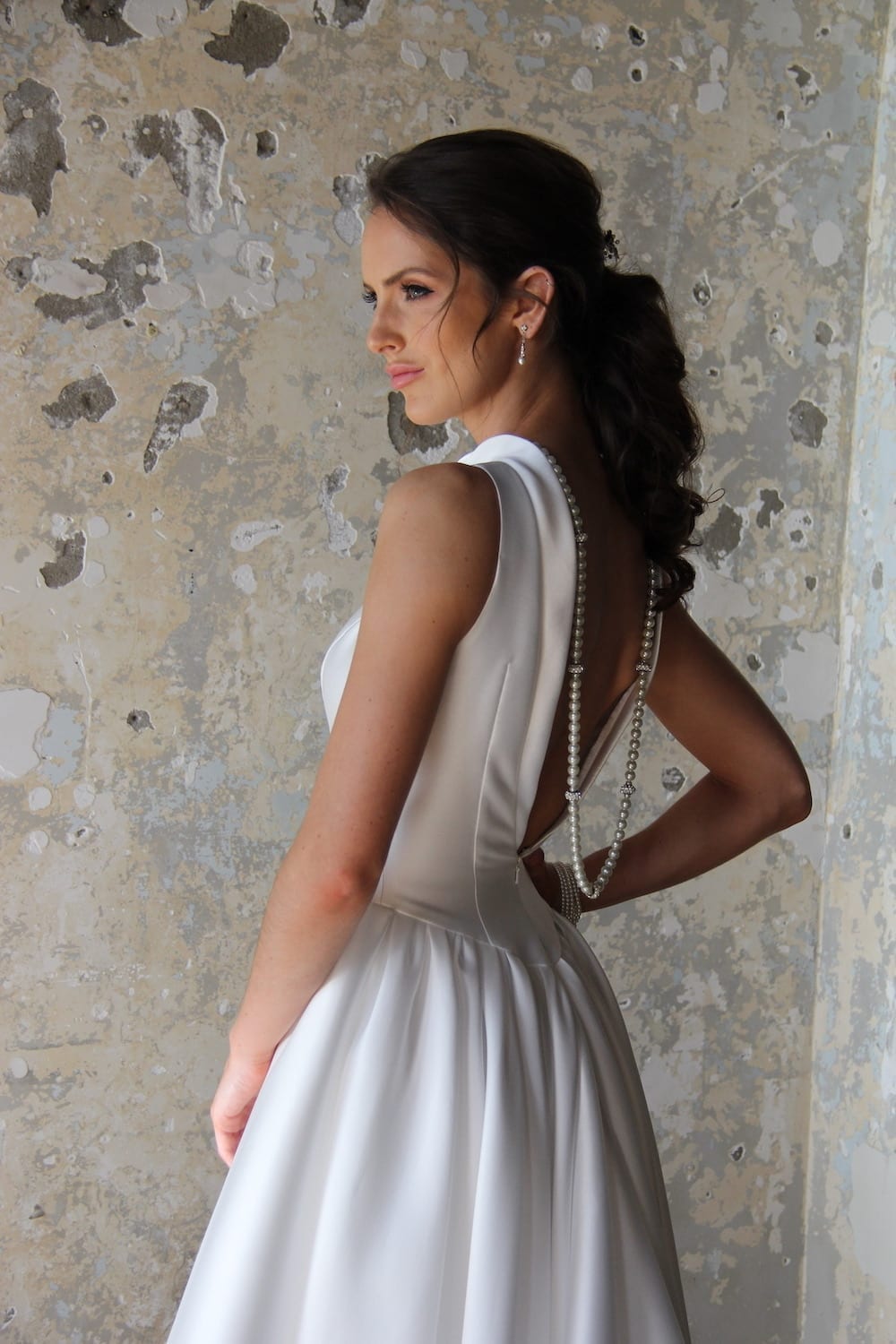 Female model wearing Vinka Design Modern Muse Wedding Dress. In chic warehouse the side detail of a gown with a high neckline and collar into a low back with a cinched waist and full Mikado satin skirt.