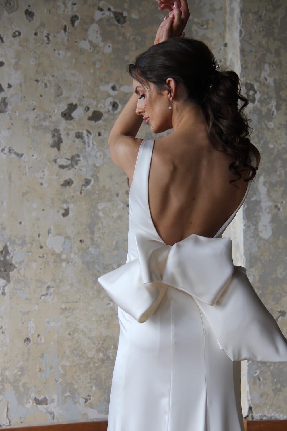 Female model wearing Vinka Design Modern Muse Wedding Dress. In chic warehouse the back detail of a gown with a bateau neckline and low scooped back gently flaring to a simple train with a detachable bow.