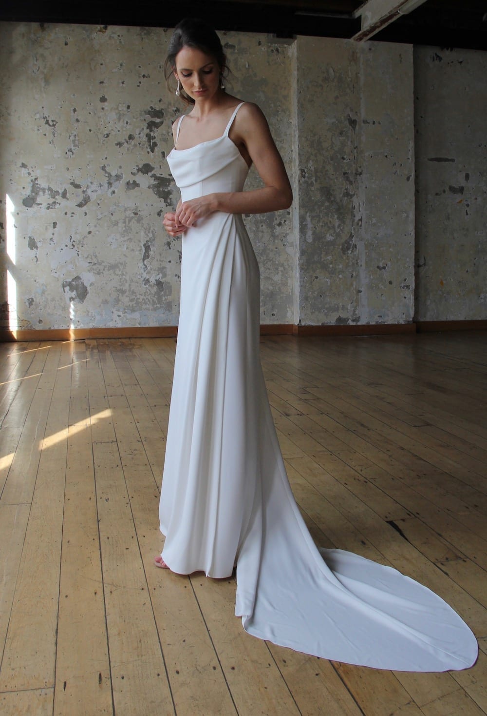 Female model wearing Vinka Design Modern Muse Wedding Dress. In chic warehouse the side detail of a gown of soft bridal crepe with a fitted bodice with soft draping over the bust line and a side split skirt.