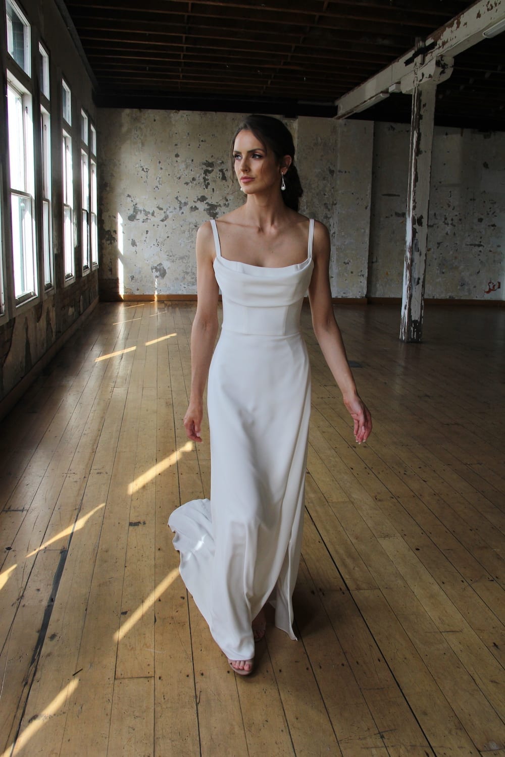 Female model wearing Vinka Design Modern Muse Wedding Dress. In chic warehouse the front detail of a gown of soft bridal crepe with a fitted bodice with soft draping over the bust line and a side split skirt.
