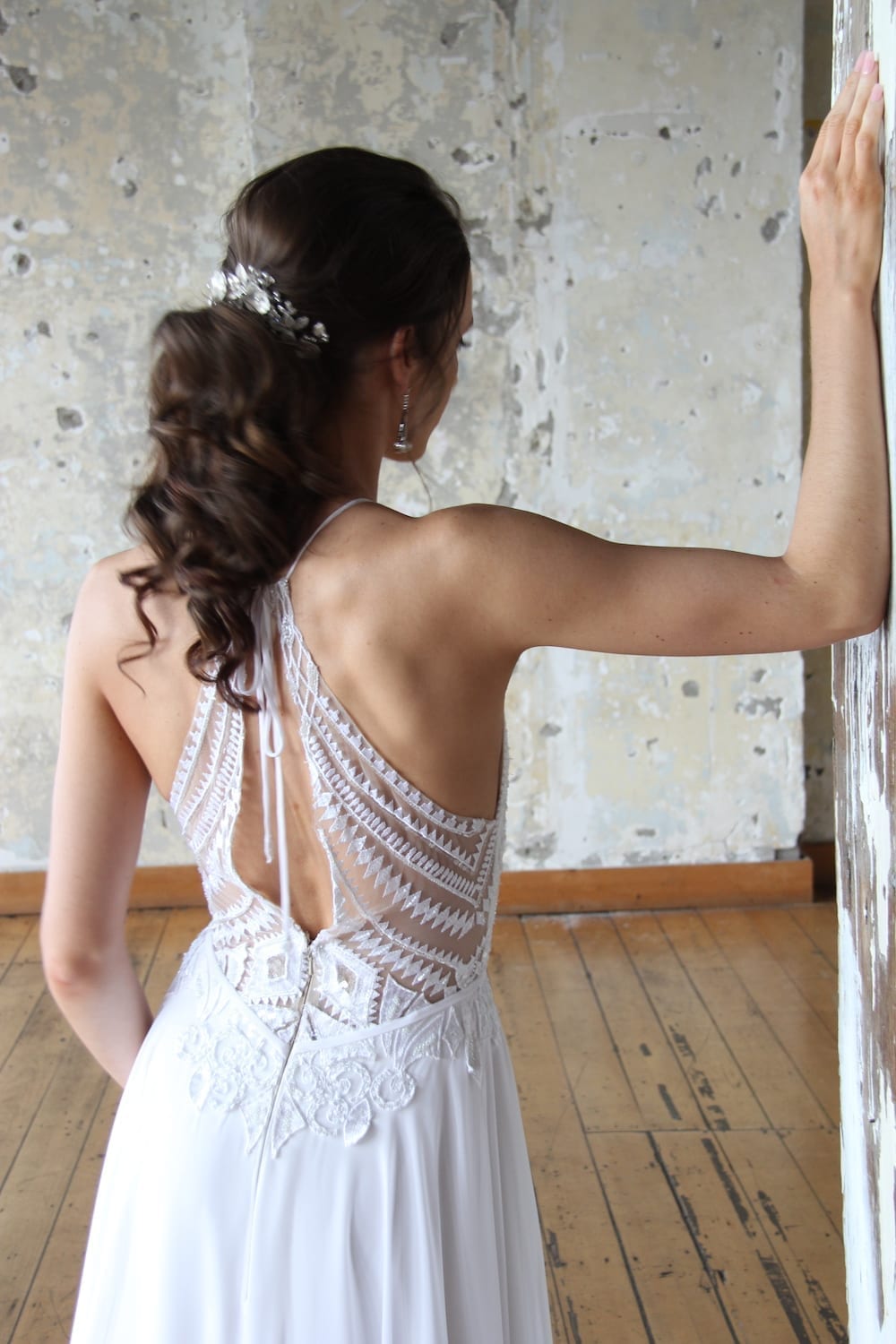 Female model wearing Vinka Design Modern Muse Wedding Dress. In chic warehouse the back detail of a light, halter-necked gown with a fluid chiffon skirt.
