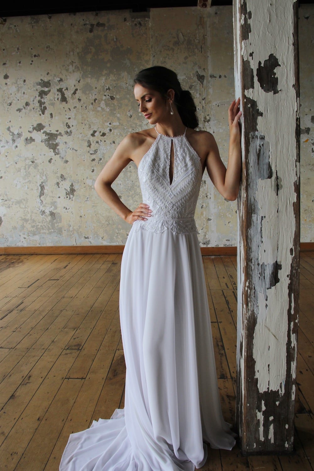 Female model wearing Vinka Design Modern Muse Wedding Dress. In chic warehouse the front detail of a light, halter-necked gown with a fluid chiffon skirt.