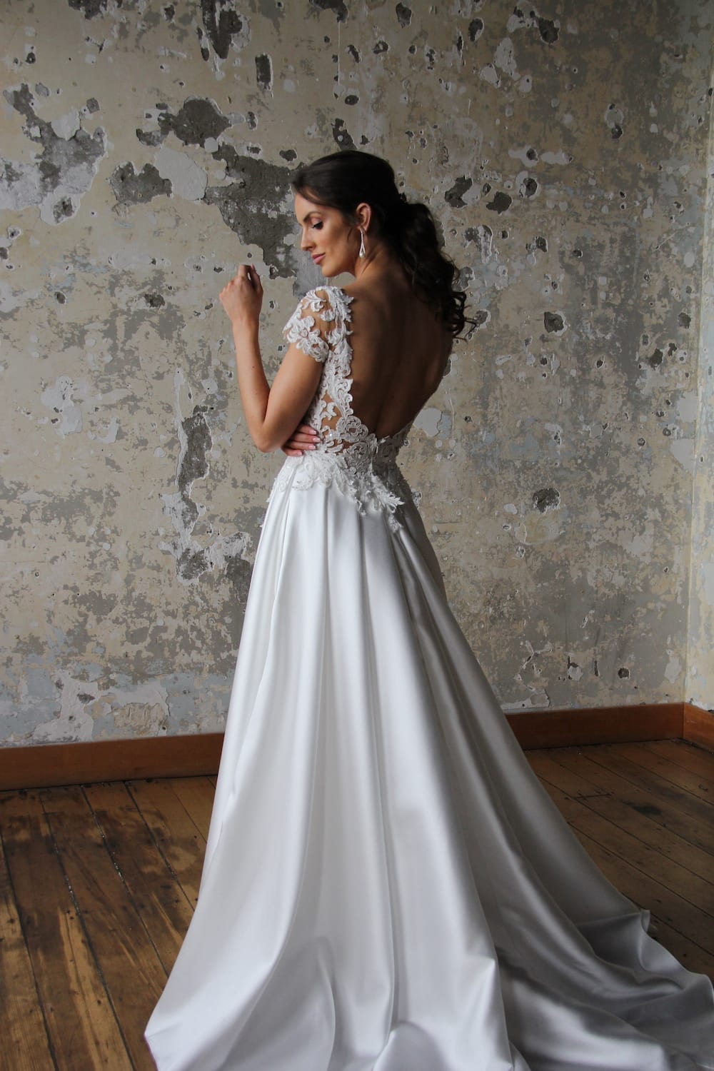 Female model wearing Vinka Design Modern Muse Wedding Dress. In chic warehouse the back detail of a gown with a sheer high neckline, low back edged with lace and a skirt with pockets.