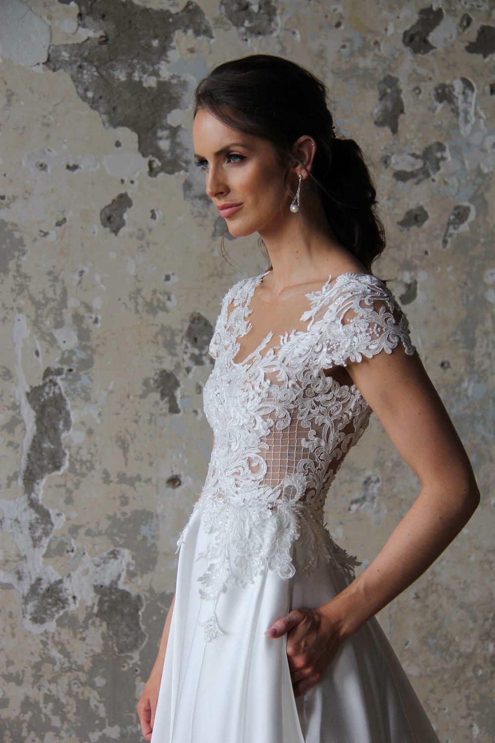 Female model wearing Vinka Design Modern Muse Wedding Dress. In chic warehouse the side detail of a gown with a sheer high neckline, low back edged with lace and a skirt with pockets.