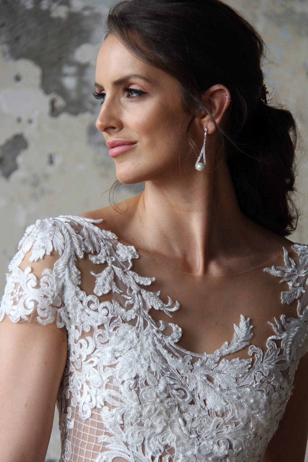 Female model wearing Vinka Design Modern Muse Wedding Dress. In chic warehouse the front detail of a gown with a sheer high neckline, low back edged with lace and a skirt with pockets.