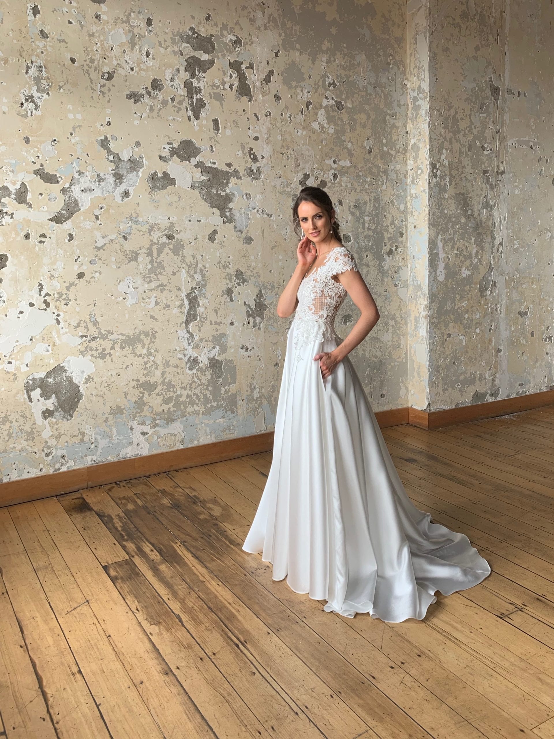Female model wearing Vinka Design Modern Muse Wedding Dress. In chic warehouse the front detail of a gown with a sheer high neckline, low back edged with lace and a skirt with pockets.