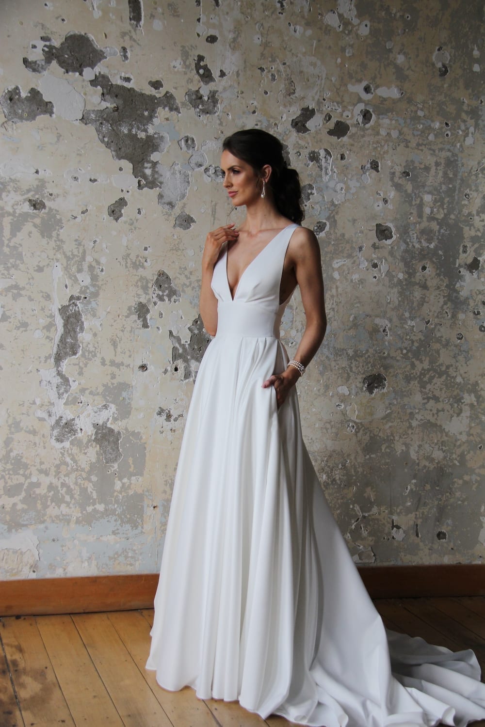 Female model wearing Vinka Design Modern Muse Wedding Dress. In chic warehouse the front detail of a gown with plunging v-neckline, Clean, sharp lines and skirt with deep pleated folds and pockets.