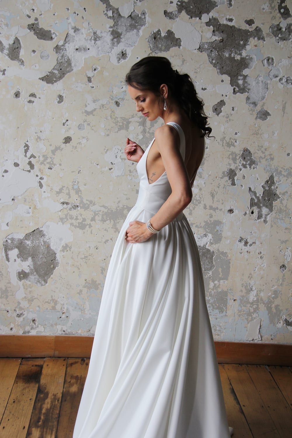 Female model wearing Vinka Design Modern Muse Wedding Dress. In chic warehouse the side detail of a gown with plunging v-neckline, Clean, sharp lines and skirt with deep pleated folds and pockets.