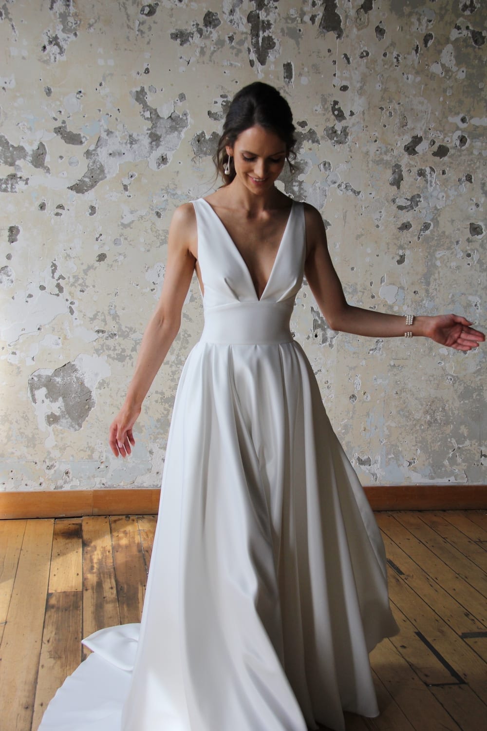 Female model wearing Vinka Design Modern Muse Wedding Dress. In chic warehouse the front detail of a gown with plunging v-neckline, Clean, sharp lines and skirt with deep pleated folds and pockets.