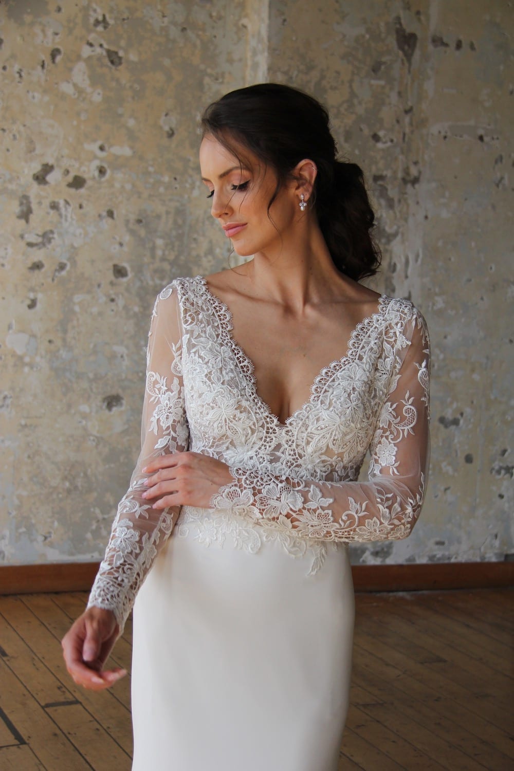 Female model wearing Vinka Design Modern Muse Wedding Dress. In chic warehouse the front detail of a gown with v-neckline, intricate bodice, long lace sleeves, and Bridal crepe skirt.