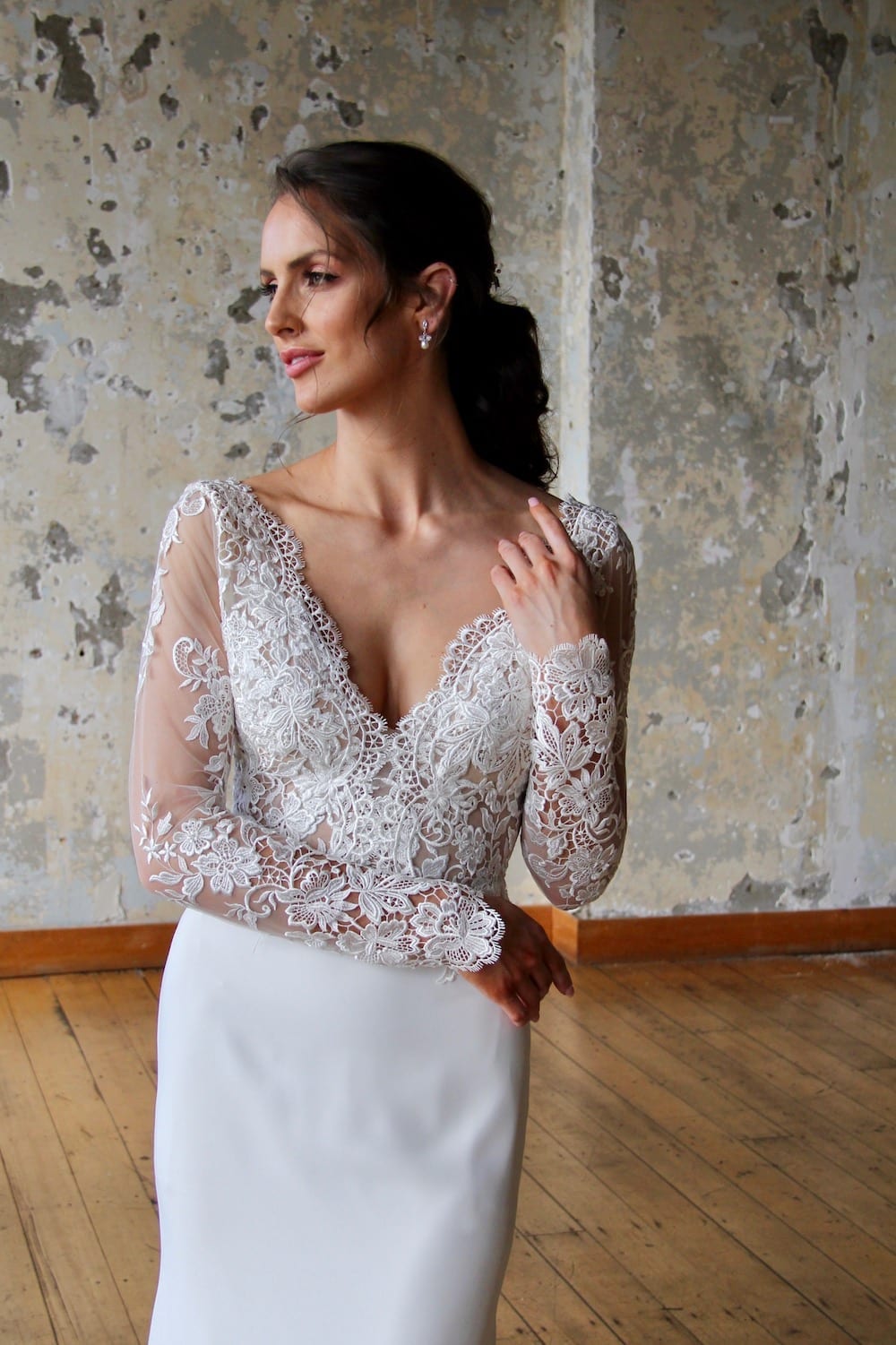 Female model wearing Vinka Design Modern Muse Wedding Dress. In chic warehouse the front detail of a gown with v-neckline, intricate bodice, long lace sleeves, and Bridal crepe skirt.