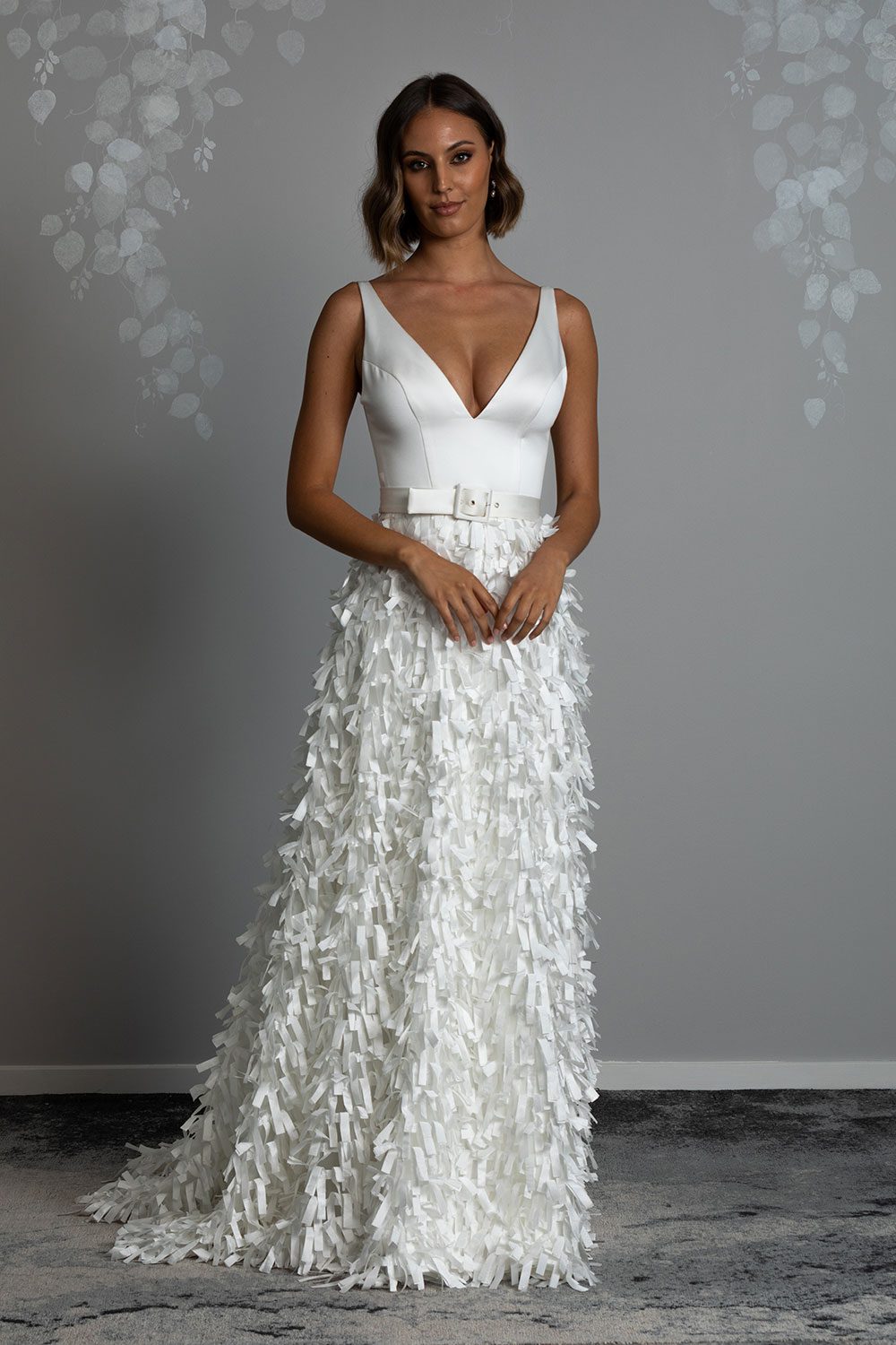 Bianca wedding dress from Vinka design. Full length view of model with hands clasped in front of belt that cinches the waist of deep V cut dress with ribbon cut skirt
