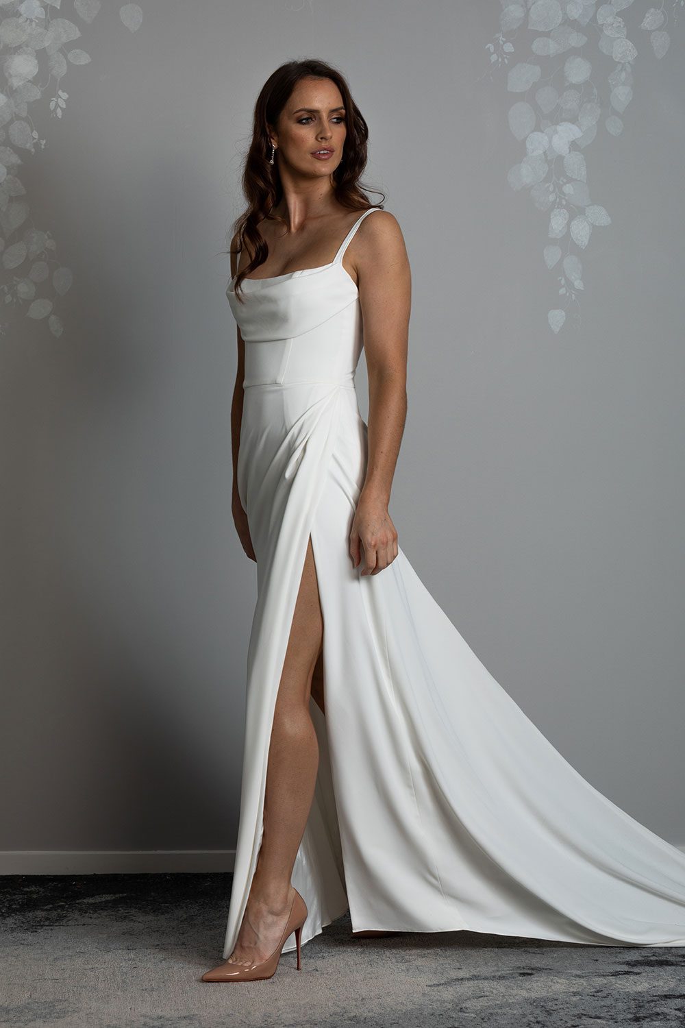 Evelyn Wedding Dress from Vinka Design. A divine gown made in a soft bridal crepe. It has a fitted bodice with soft draping over the bust line to flatter and enhance the figure. The straps compliment this detail by giving the bodice a softened square neckline. The skirt elegantly falls in folds from the side, revealing a flirtatious side split while in motion that falls closed again when still. Model showing side split in soft bridal crepe skirt