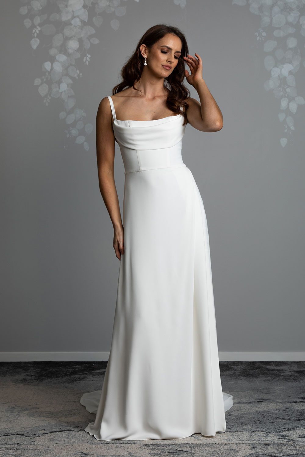 Evelyn Wedding Dress from Vinka Design. A divine gown made in a soft bridal crepe. It has a fitted bodice with soft draping over the bust line to flatter and enhance the figure. The straps compliment this detail by giving the bodice a softened square neckline. The skirt elegantly falls in folds from the side, revealing a flirtatious side split while in motion that falls closed again when still. Model with hand to face wearing softly draped fitted bodice with bridal crepe skirt