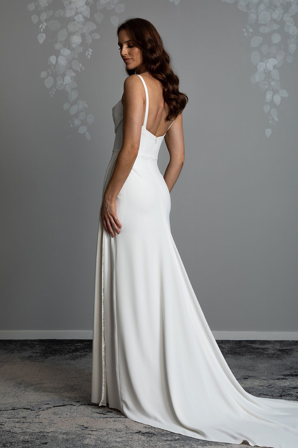 Evelyn Wedding Dress from Vinka Design. A divine gown made in a soft bridal crepe. It has a fitted bodice with soft draping over the bust line to flatter and enhance the figure. The straps compliment this detail by giving the bodice a softened square neckline. The skirt elegantly falls in folds from the side, revealing a flirtatious side split while in motion that falls closed again when still. Back view of long bridal crepe train and flattering square back design