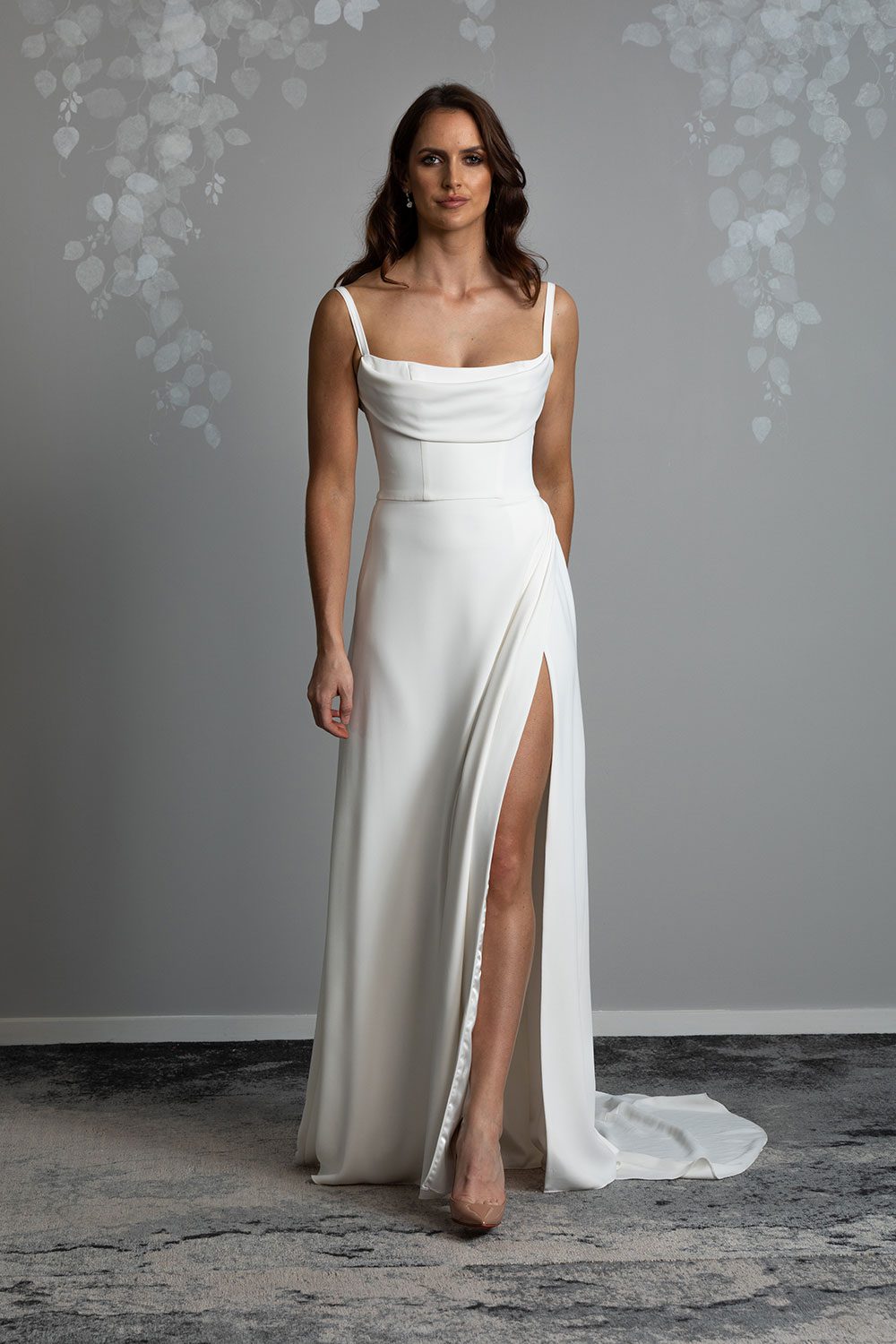 Evelyn Wedding Dress from Vinka Design. A divine gown made in a soft bridal crepe. It has a fitted bodice with soft draping over the bust line to flatter and enhance the figure. The straps compliment this detail by giving the bodice a softened square neckline. The skirt elegantly falls in folds from the side, revealing a flirtatious side split while in motion that falls closed again when still. Stunning front view of model walking in gown with fitted bodice with draping and long bridal crepe skirt with side split