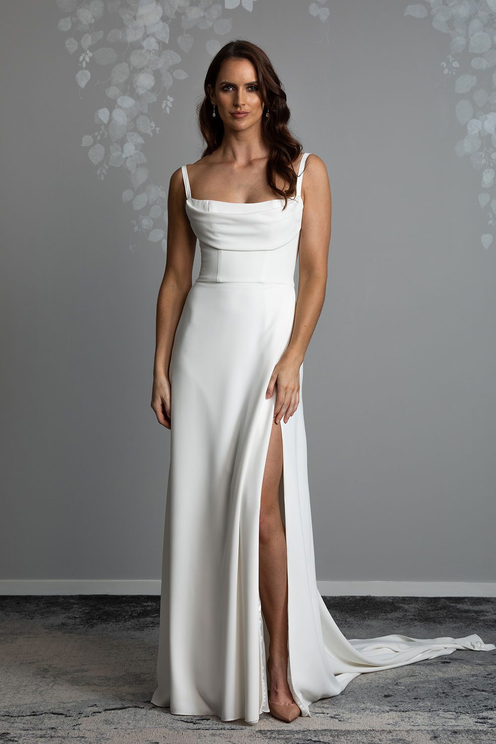 Evelyn Wedding Dress from Vinka Design. A divine gown made in a soft bridal crepe. It has a fitted bodice with soft draping over the bust line to flatter and enhance the figure. The straps compliment this detail by giving the bodice a softened square neckline. The skirt elegantly falls in folds from the side, revealing a flirtatious side split while in motion that falls closed again when still. Full length view of model wearing stunning gown with fitted bodice with draping at the bust and long bridal crepe skirt with side split and lengthy train
