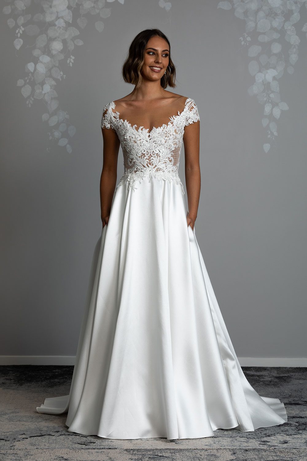 Leona Wedding Dress from Vinka Design. This gorgeous gown is hand appliqued with two complimentary laces on a nude tulle base, which creates a striking effect. Delicate cap sleeves flatter the arms whilst also drawing the eye to the slenderness of the waist. It has a low back edged with lace that cinches in the waist and is carefully styled to delicately finish into the dramatic skirt. Full length view of model wearing with intricate lace bodice and cap sleeves with silk skirt that cinches at the waist and long sillk skirt with folds and pockets