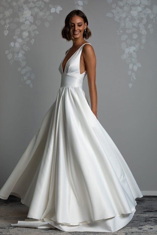Roma Wedding Dress from Vinka Design. Gown with plunging v-neckline, Clean, sharp lines and skirt with deep pleated folds and pockets. Model spinning sideways wearing plunging V neckline dress fitted to a yolk with Ottoman satin skirt that shows beautiful movement in the deep folds