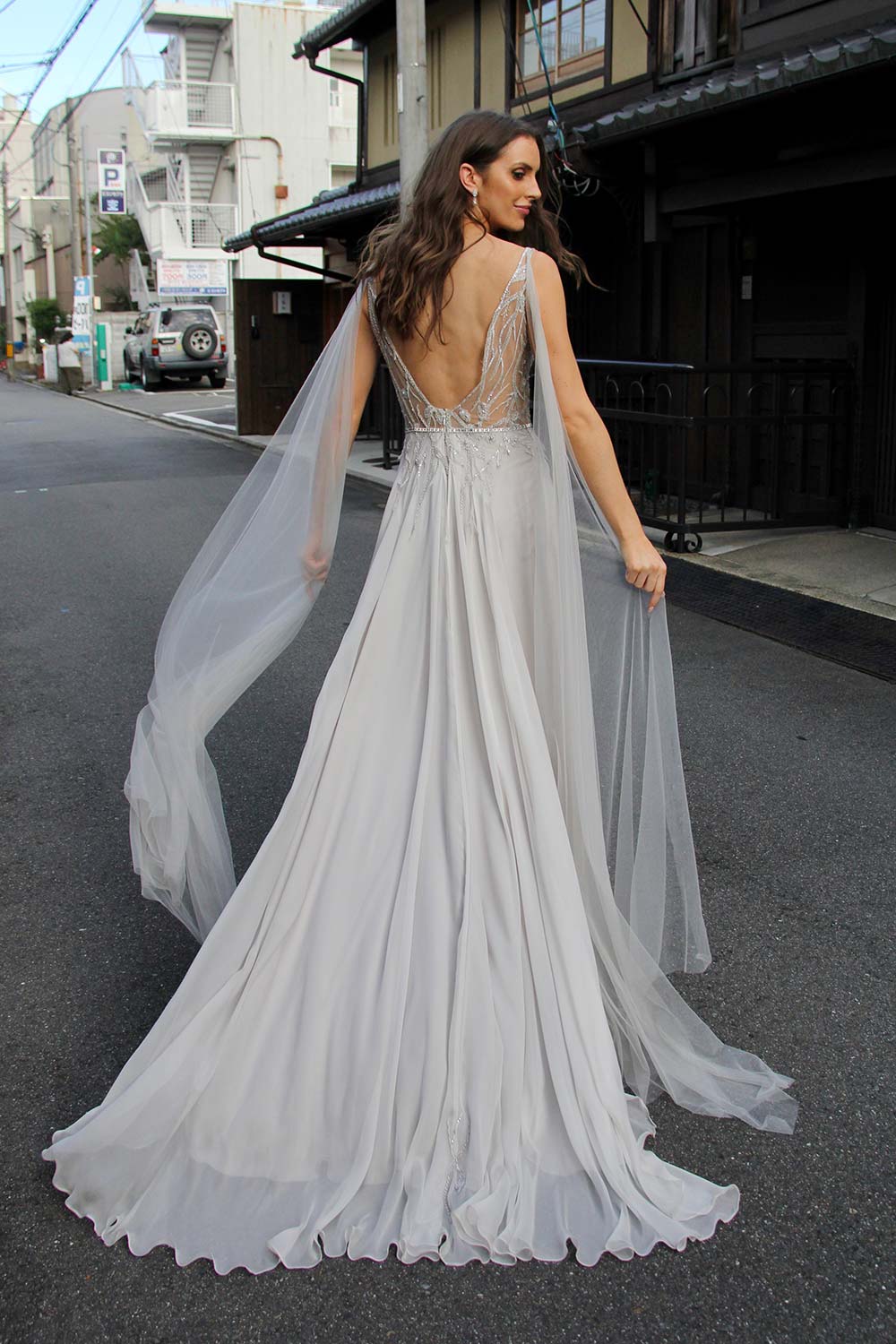 Female model wearing Vinka Design An Oriental Affair Wedding Dress. On a Japanese street the back detail of a gown with a sheer, deep v-neckline bodice with swarovski diamante belt, tulle cape and silver silk chiffon skirt.