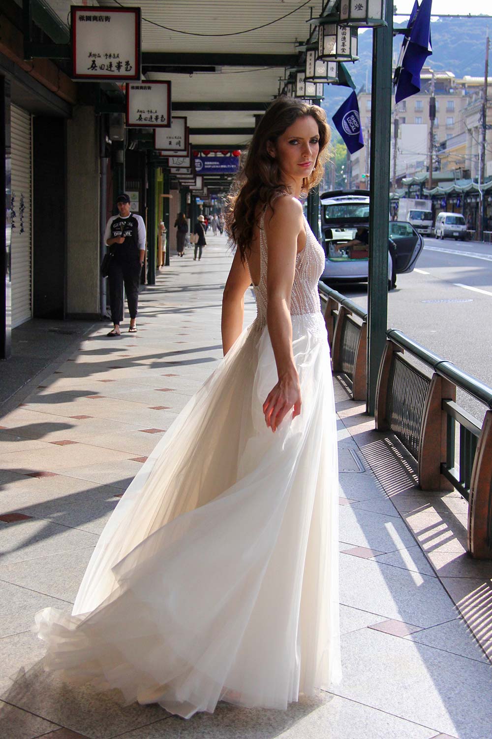 Female model wearing Vinka Design An Oriental Affair Wedding Dress. On a Japanese street the side detail of a gown with a hand-beaded, nude-based, semi sheer bodice with a deep v-neckline and low back with a tulle skirt.