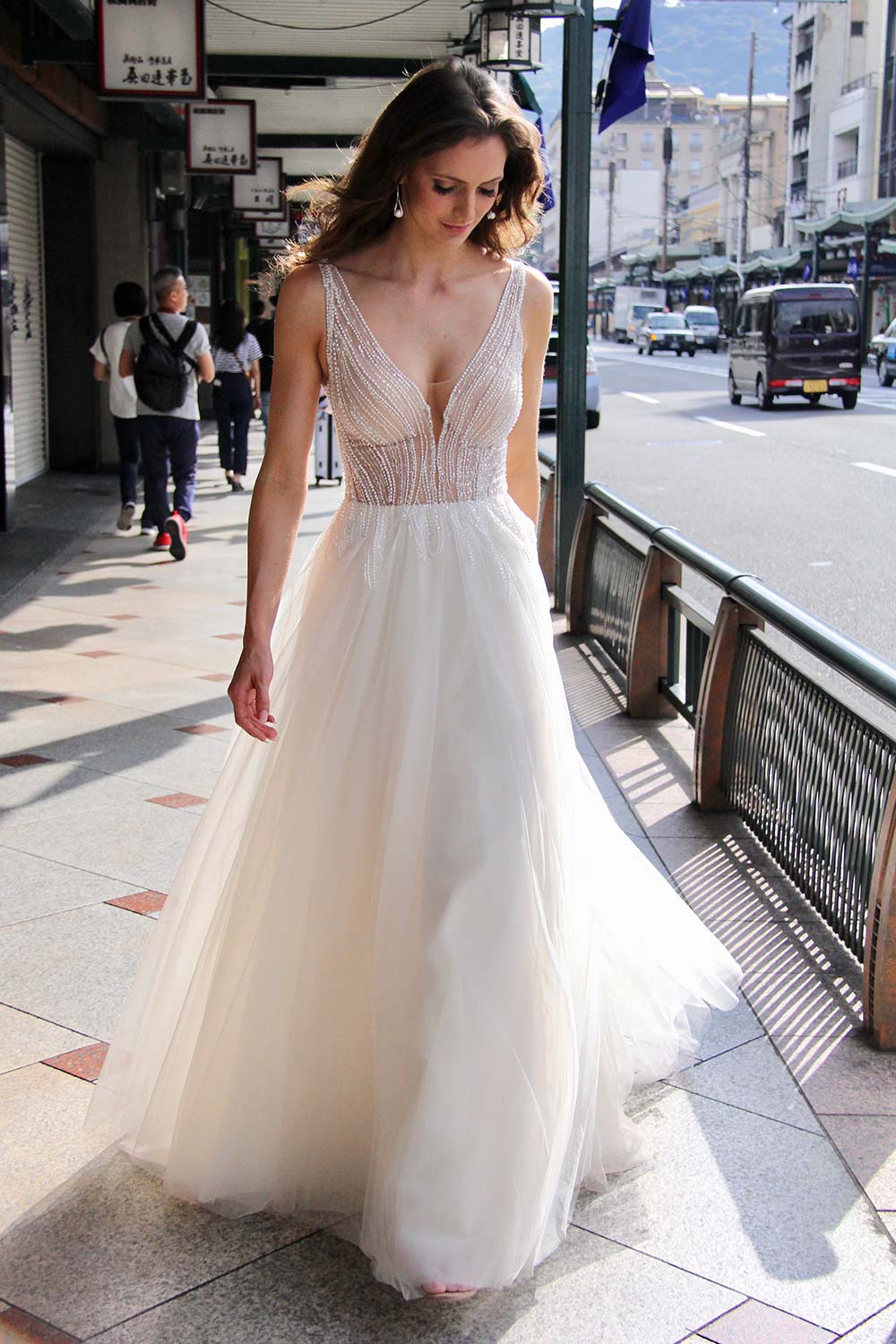 Female model wearing Vinka Design An Oriental Affair Wedding Dress. On a Japanese street the back detail of a gown with a hand-beaded, nude-based, semi sheer bodice with a deep v-neckline and low back with a tulle skirt.
