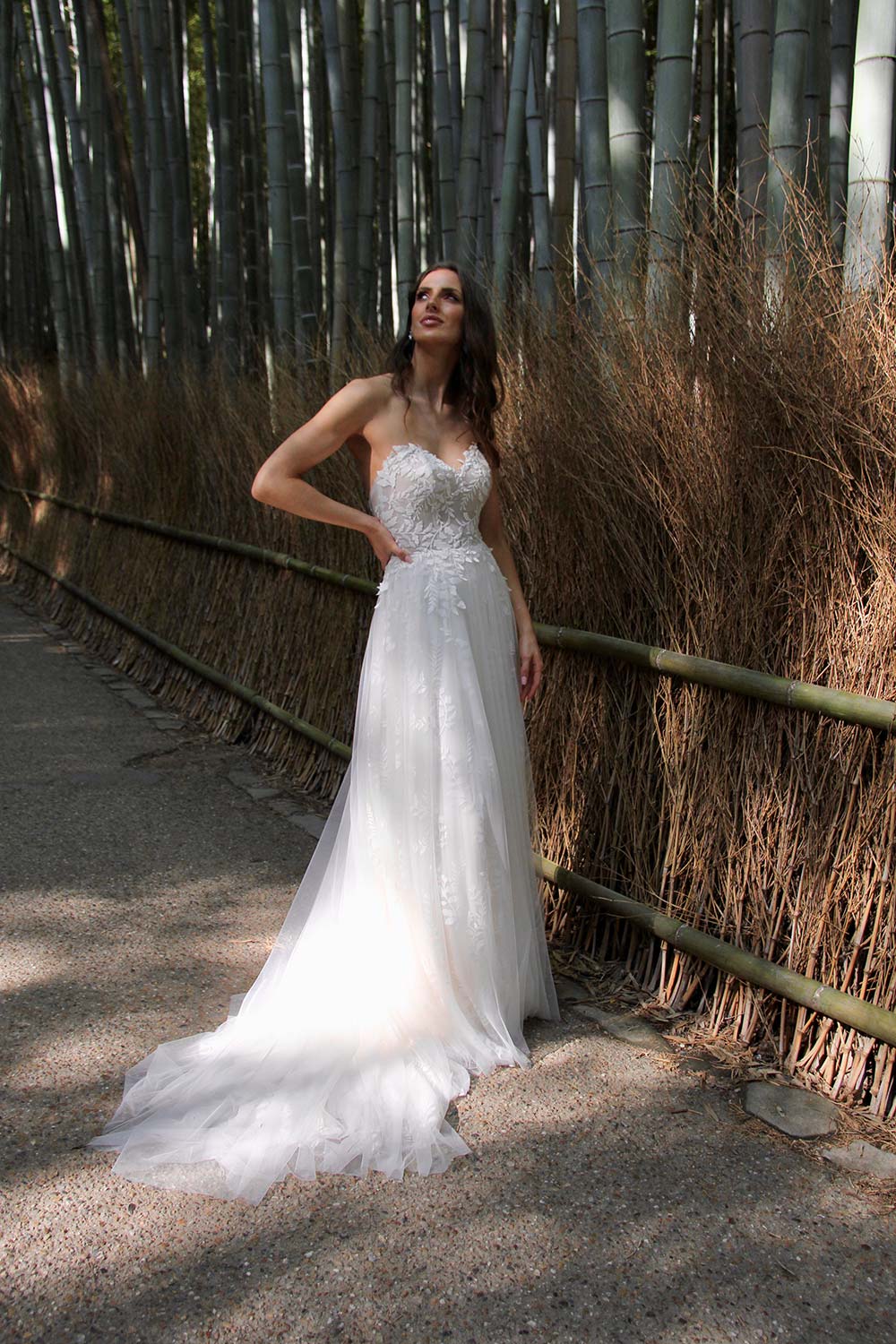 Female model wearing Vinka Design An Oriental Affair Wedding Dress. In a Japanese bamboo forest the front detail of gown with a strapless bodice with leaf lace and a full length train skirt