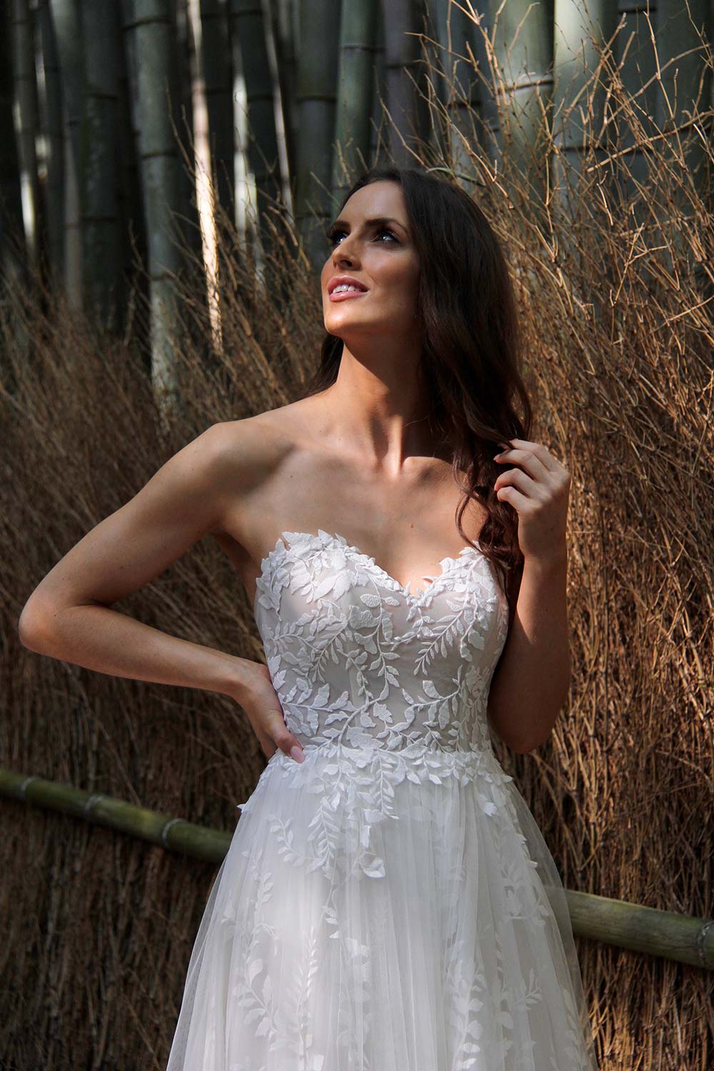 Female model wearing Vinka Design An Oriental Affair Wedding Dress. In a Japanese bamboo forest the front detail of gown with a strapless bodice with leaf lace and a full length train skirt