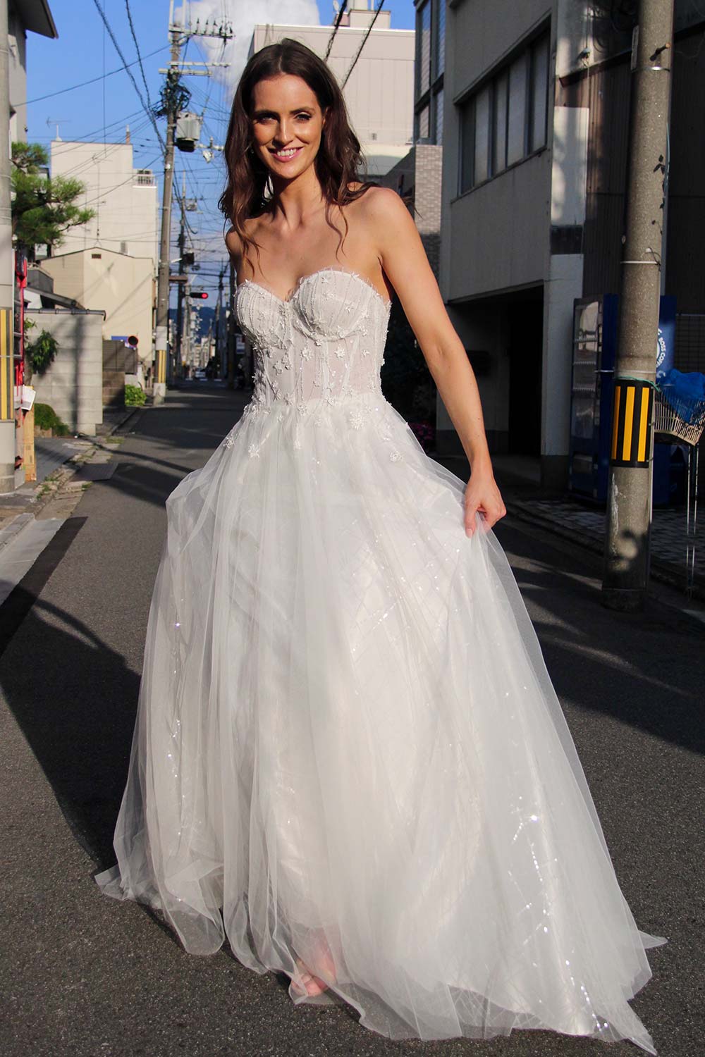 Female model wearing Vinka Design An Oriental Affair Wedding Dress. On a japanese street the front detail of a semi-sheer bodice with boning and hand-appliqued lace and skirt made with multiple layers of tulle
