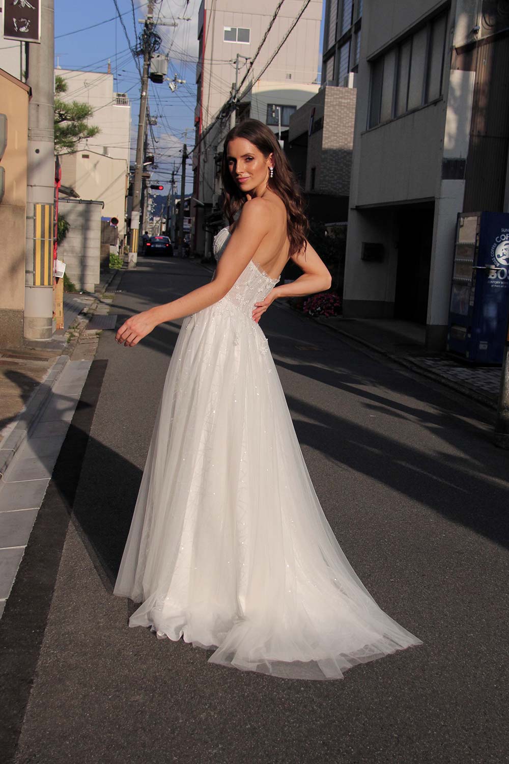 Female model wearing Vinka Design An Oriental Affair Wedding Dress. On a japanese street the side detail of a semi-sheer bodice with boning and hand-appliqued lace and skirt made with multiple layers of tulle