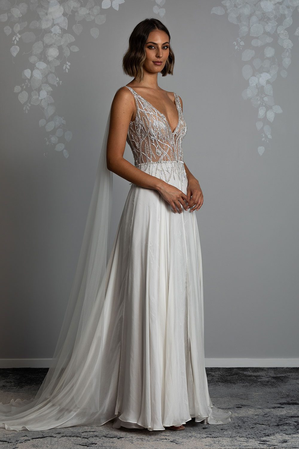 Amaya Wedding Dress from Vinka Design. This stunning gown is made with a sheer, deep v-neckline both front and back, with richly beaded silver lace appliqued throughout the bodice. The bodice is then tied together with an adorning swarovski diamante belt and tulle cape trailing from the shoulder, which can be made detachable. The skirt consists of flowing silver silk chiffon, which opens to reveal lace applique purposefully placed at the front of the skirt. Semi profile view of model with sheer deep V-neckline and flowing silver silk chiffon skirt and tulle cape