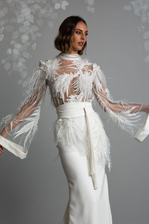 Miu Wedding Dress from Vinka Design. The bodice is made with a dimensional beaded lace adorned with sequins and feathers. The high neckline compliments the gorgeous kimono-like sleeves, with hand-stitched bridal crepe cuffs. Model showcasing the kimono sleeves with feathered and beaded details, and wide belt that cinches the waist