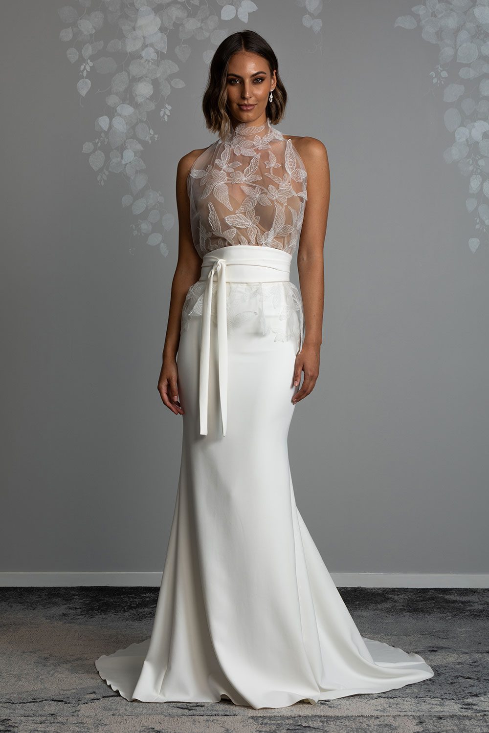 Mio Wedding Dress from Vinka Design. The front detail of gown with a high necked delicately hand-embroidered leaf lace cinched in at the waist with a wide kimono belt with skirt. Model wearing high necked leaf lace tulle bodice with kimono belt, and long skirt with train
