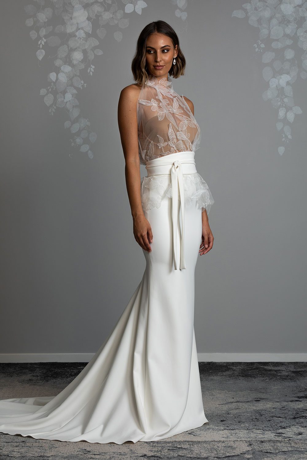 Mio Wedding Dress from Vinka Design. The front detail of gown with a high necked delicately hand-embroidered leaf lace cinched in at the waist with a wide kimono belt with skirt. Model wearing high necked leaf lace tulle bodice with kimono belt, and skirt with long train